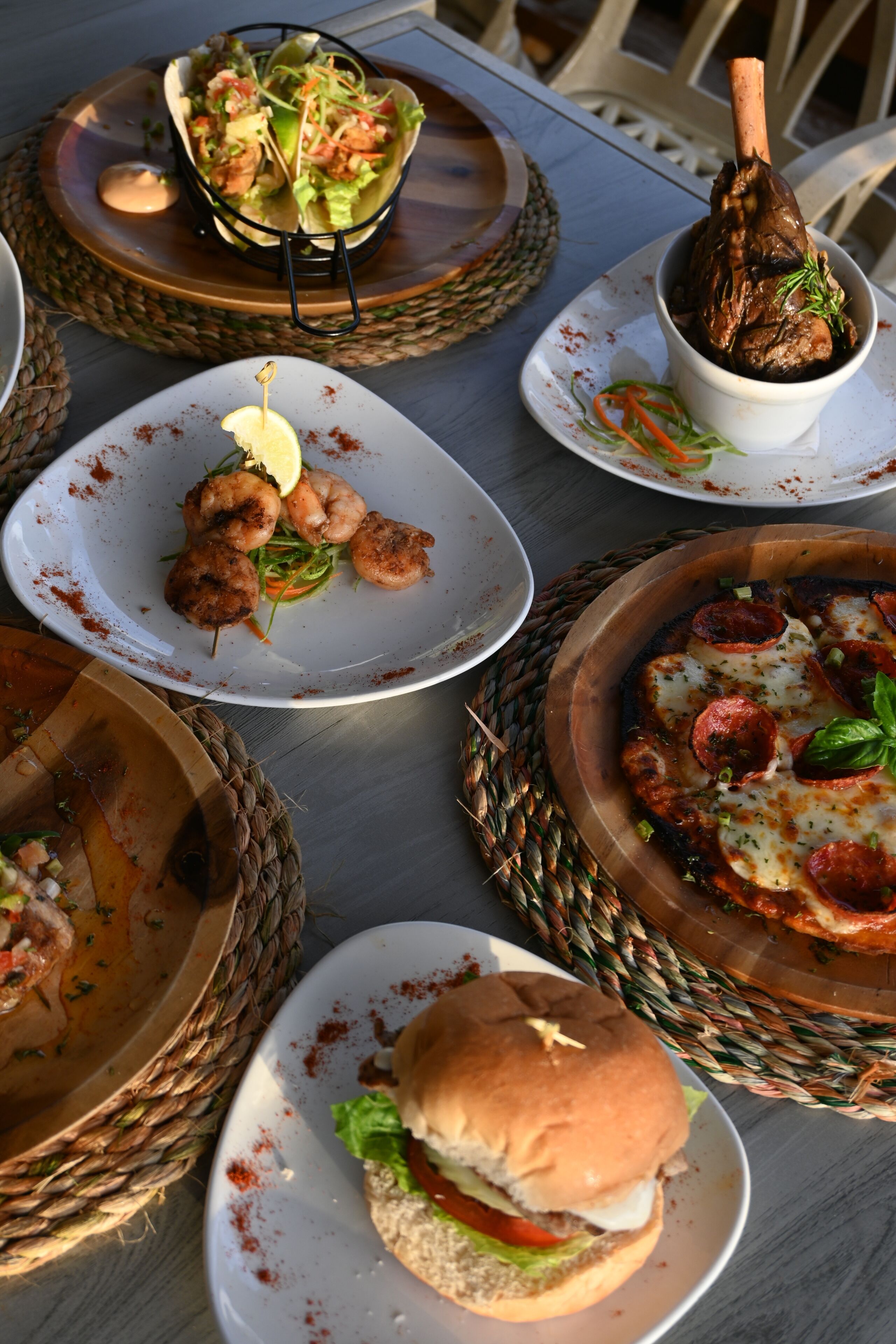 Una mesa servida con diversos platillos, incluyendo tacos, camarones, una hamburguesa y una pizza.