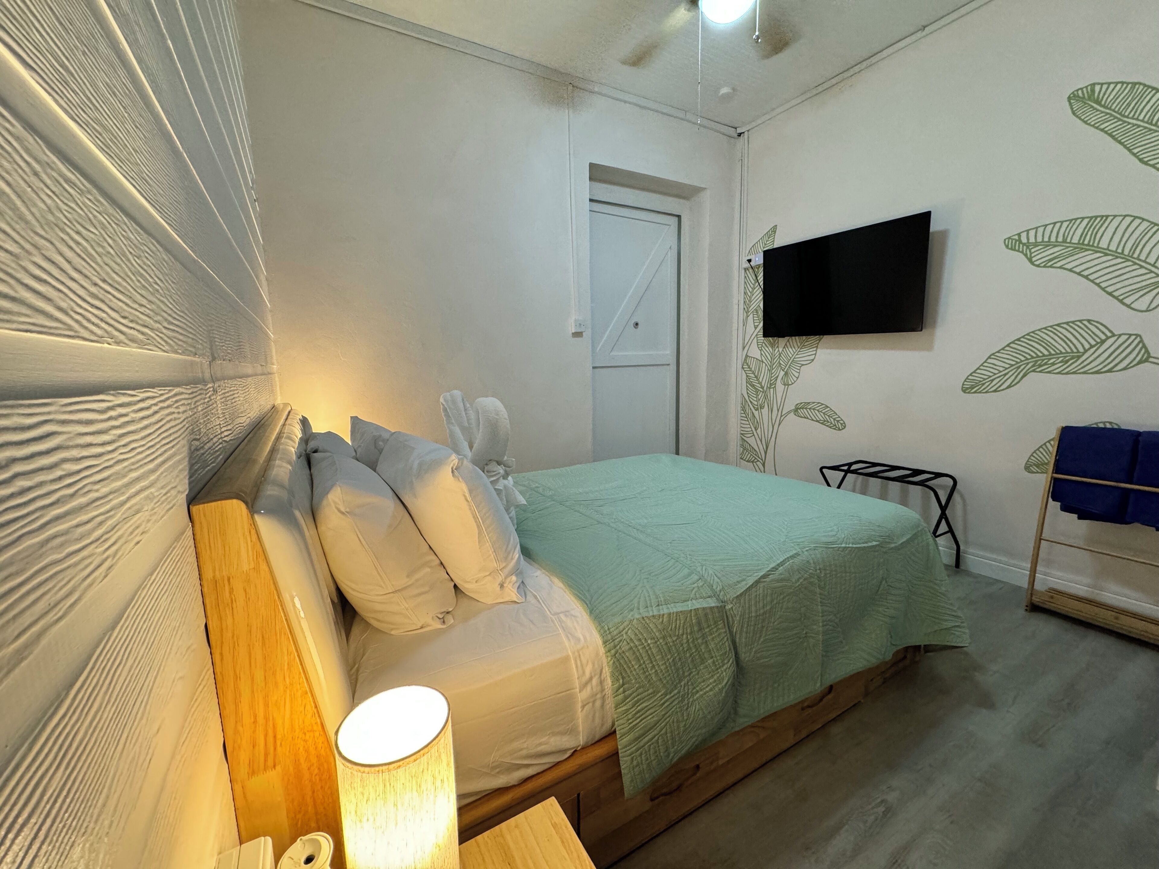 A bedroom with a bed, pillows, a bedside lamp, a TV on the wall, and a towel rack.