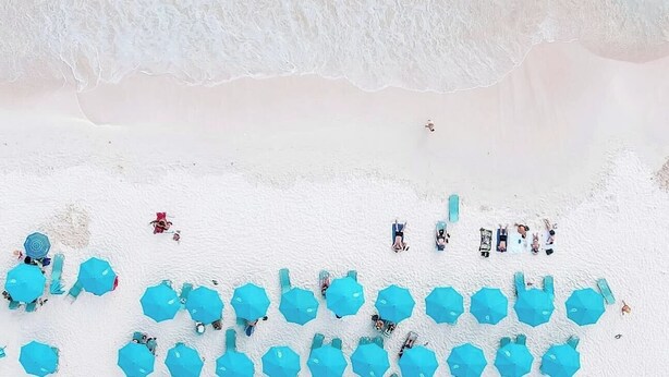 Een luchtend vanuit de lucht zicht op een strand met turquoise water, wit zand en rijen met blauwe parasols.