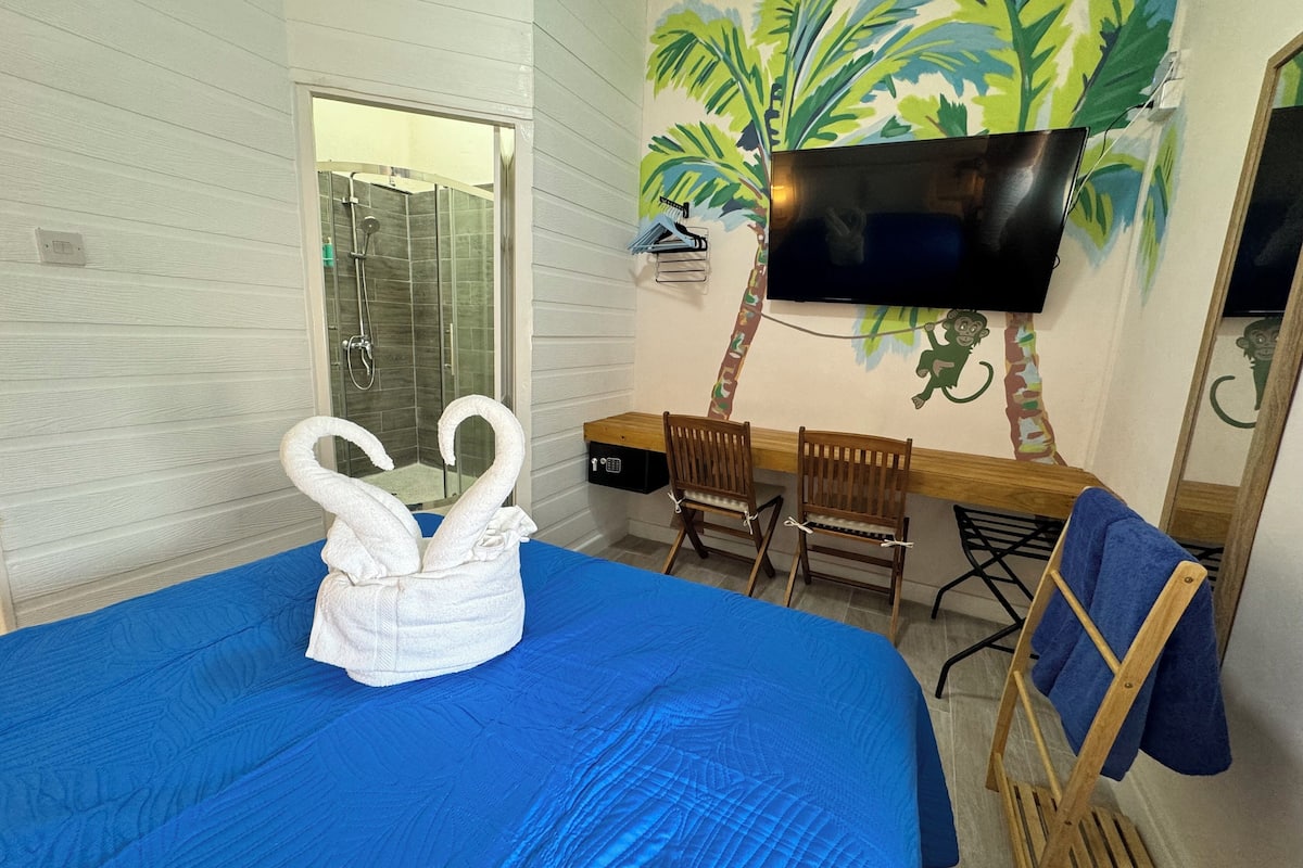 A hotel room with a large bed, a shower, a desk with chairs, a TV mounted on the wall, and a mural of palm trees.