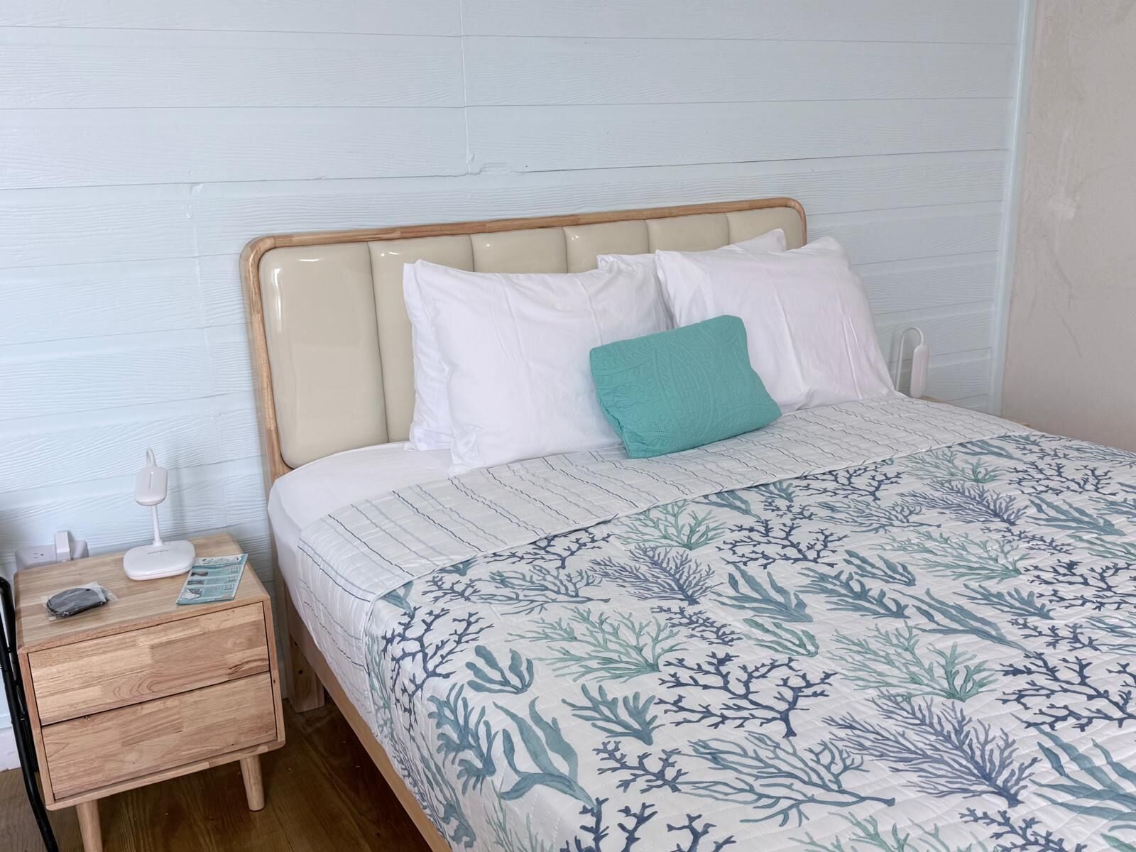 Een net opgemaakt bed met een bedsprei met patroon, een houten nachtkastje met een lamp en een teal-kleurig kussen.