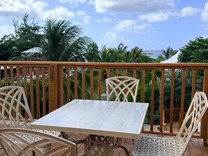 A wooden deck with a table and chairs, overlooking a beach and palm trees. - Eden On The Sea (Bridgetown)