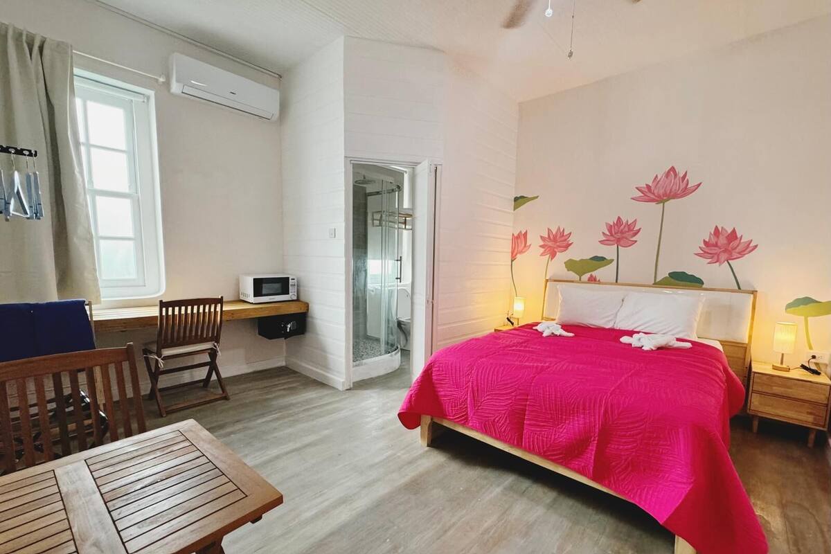 A hotel room with a bed, a desk, a chair, a microwave, a shower, and a window.