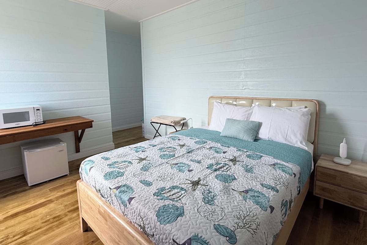 A bedroom with a wooden bed, a bedside table, a nightstand, a desk with a microwave, and a small white cabinet.