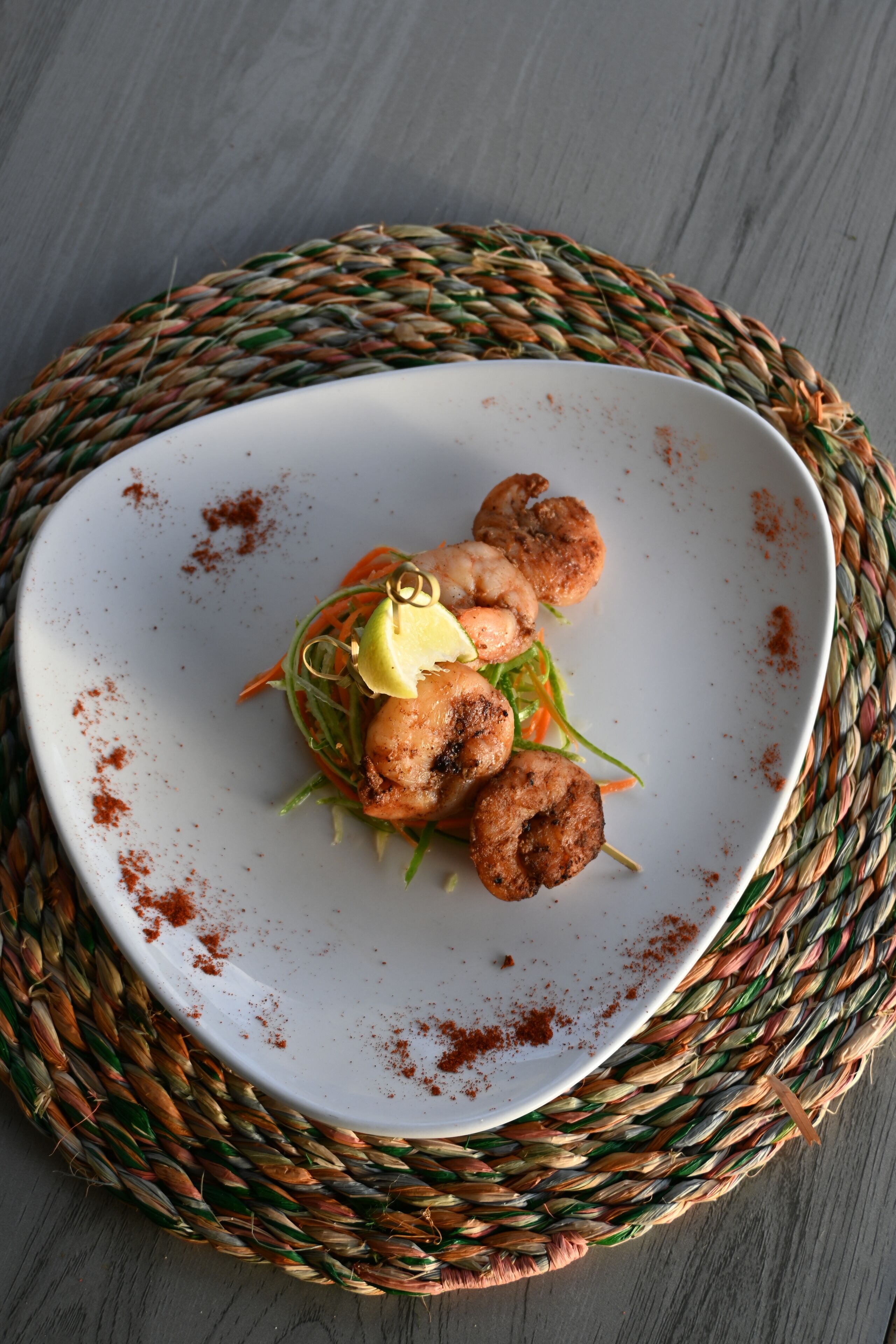 Un plato blanco con camarones a la parrilla y verduras sobre una estera tejida.