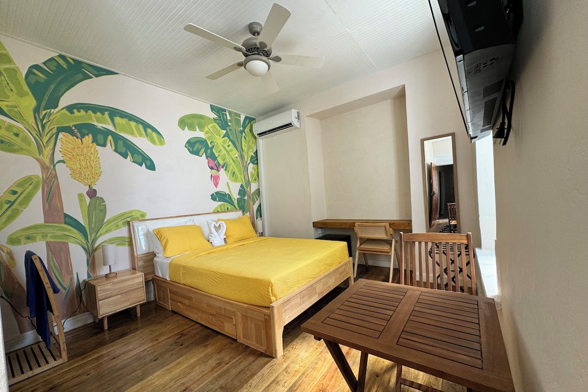 A room with a bed, a desk, a chair, a ceiling fan, and a wall-mounted TV.