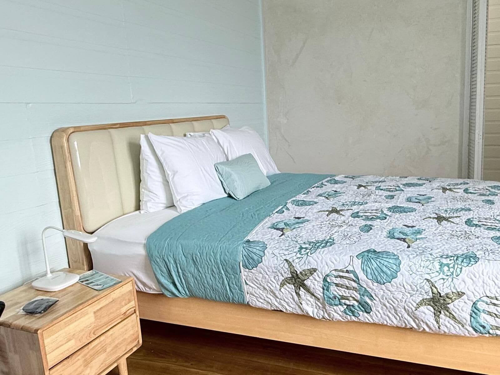 Un letto rifatto con cura, con una coperta a motivi decorativi, una testiera in legno e un comodino con una lampada e un piccolo cassetto.
