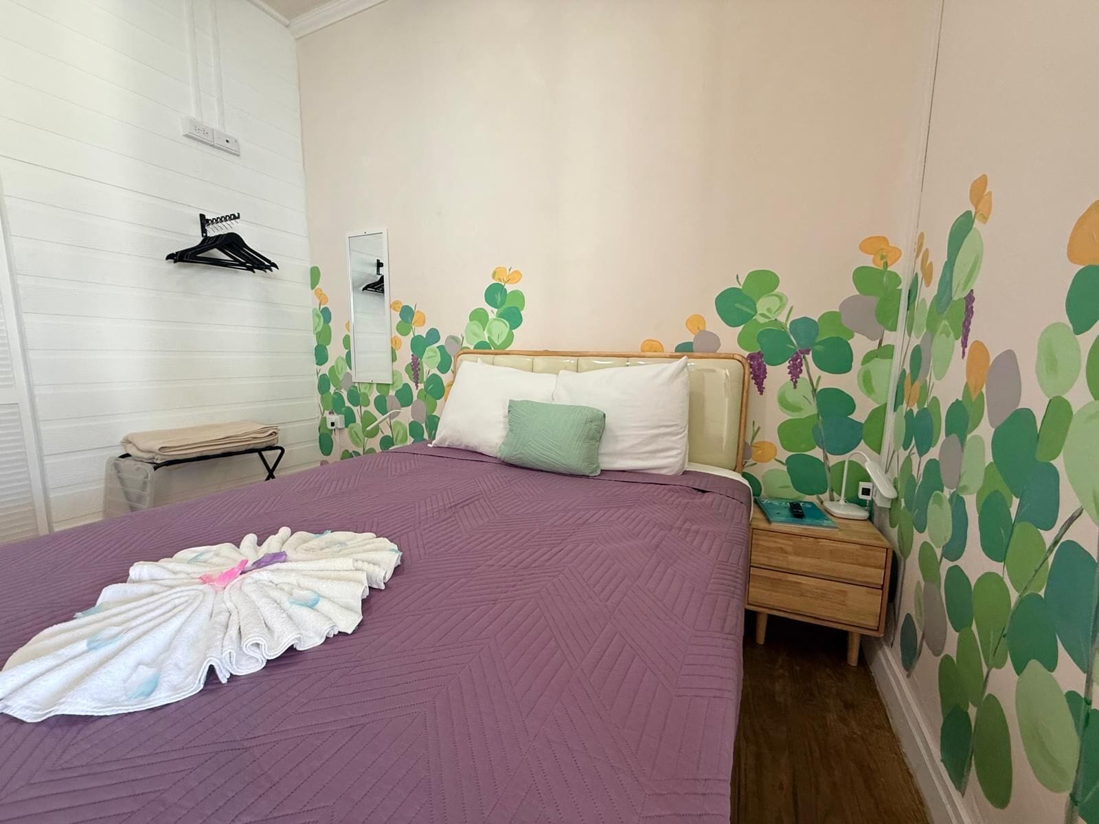 Una cama bien hecha con una colcha morada y una toalla doblada. Hay una mesita de noche con una lámpara y un cajón pequeño. La habitación tiene una pared blanca con un estampado de hojas verdes y un cabecero de madera.