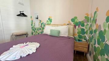 Una cama bien hecha con una colcha morada y una toalla doblada. Hay una mesita de noche con una lámpara y un cajón pequeño. La habitación tiene una pared blanca con un estampado de hojas verdes y un cabecero de madera.