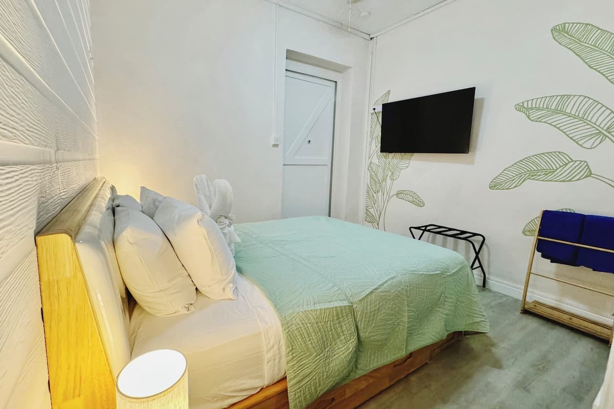 A bedroom with a bed, pillows, a bedside table with a lamp, a TV on the wall, and a wooden floor.