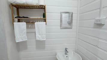 Un baño con estante de madera con toallas y artículos de aseo, un espejo pequeño y un lavabo blanco.