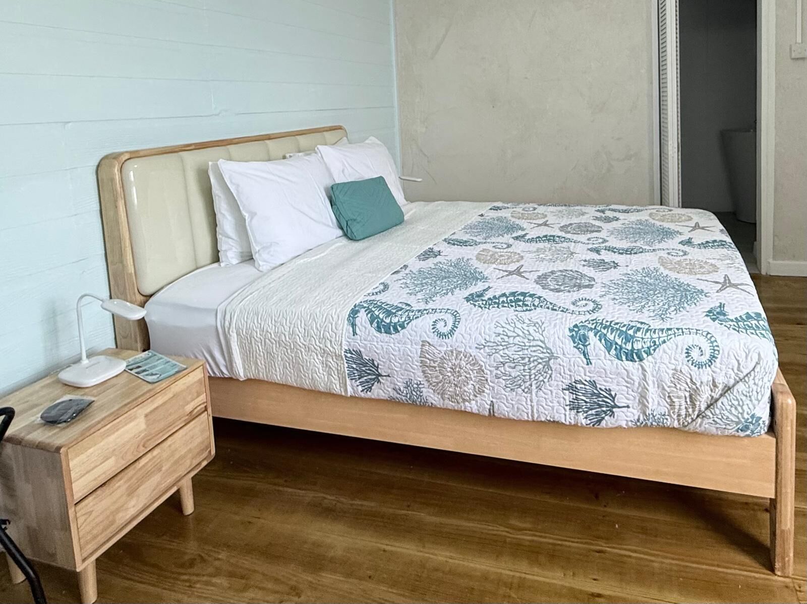 Un dormitorio con estructura de cama de madera, ropa de cama blanca, un cojín azul verdoso y una colcha estampada.
