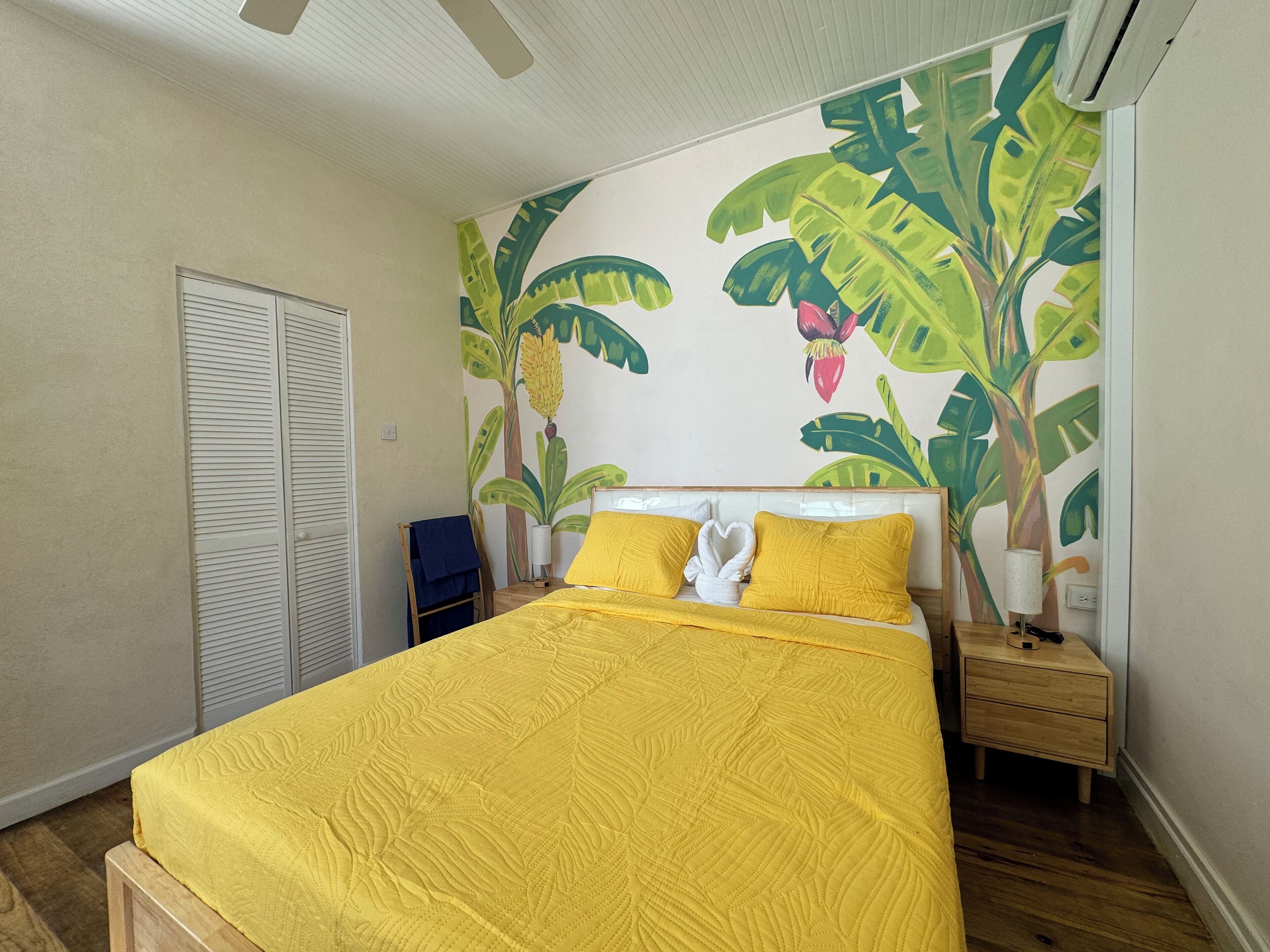 A bedroom with a bed covered in yellow bedding, a wooden nightstand with a lamp, and a wall mural of tropical leaves and flowers.