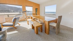 Una habitación con una gran mesa de madera, sillas, un telescopio y vistas a la playa.