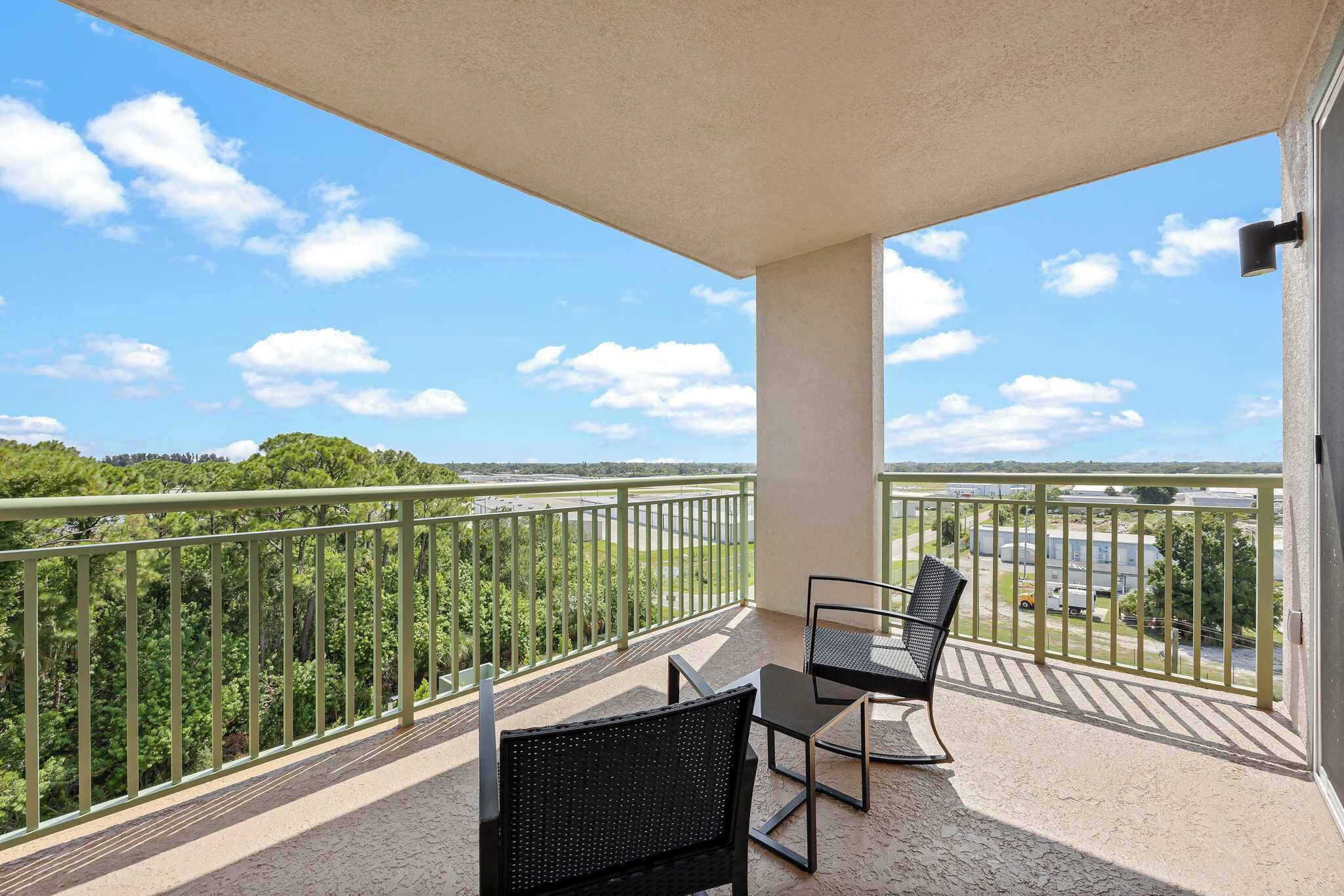 A balcony with two chairs and a table, overlooking a forested area and a distant view of a body of water.