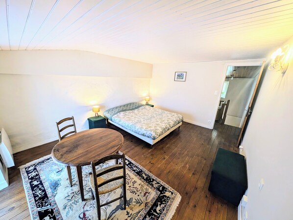 A small, cozy room with a bed, a wooden table, and a chair. There are two bedside lamps and a rug on the floor.