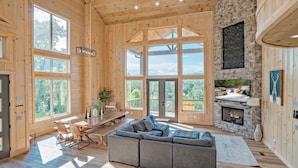 A spacious living room with a large gray sectional sofa, a stone fireplace, and large windows offering a view of the outdoors. - Casa Bella: Mtn Views, Movie Room, Games, Hot Tub, Wifi, Yard with Fire Pit! (Sevierville)