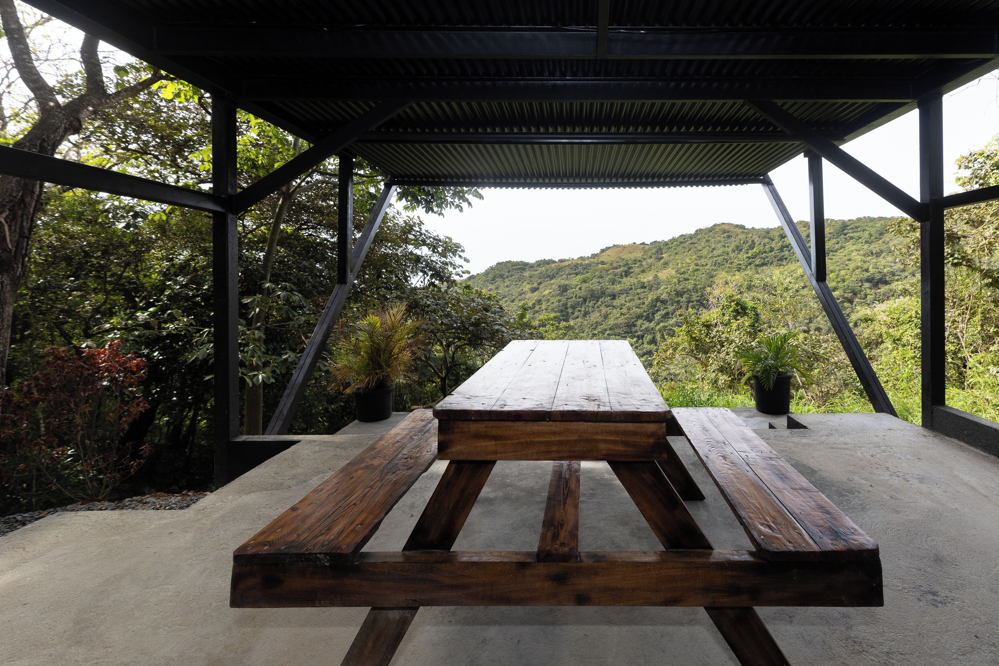 A covered outdoor seating area with a concrete floor and wooden benches, surrounded by lush greenery and a view of a hilly landscape.