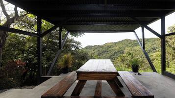 A covered outdoor seating area with a concrete floor and wooden benches, surrounded by lush greenery and a view of a hilly landscape.