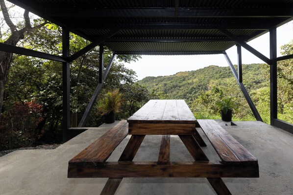 A covered outdoor seating area with a concrete floor and wooden benches, surrounded by lush greenery and a view of a hilly landscape.