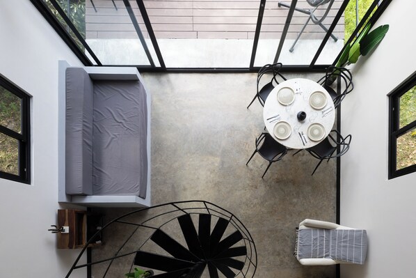 A compact room with a bed, a dining table with chairs, a spiral staircase, and a window with a view of greenery.