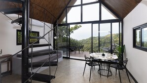 A modern interior with a staircase, a dining area with a table and chairs, and large windows offering a view of greenery and hills.