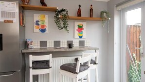 A kitchen with a built-in fridge, a counter with two white chairs, a radiator, and a window with a view of a wooden fence. - Darlington 3 bed House Near Train Station (Darlington)