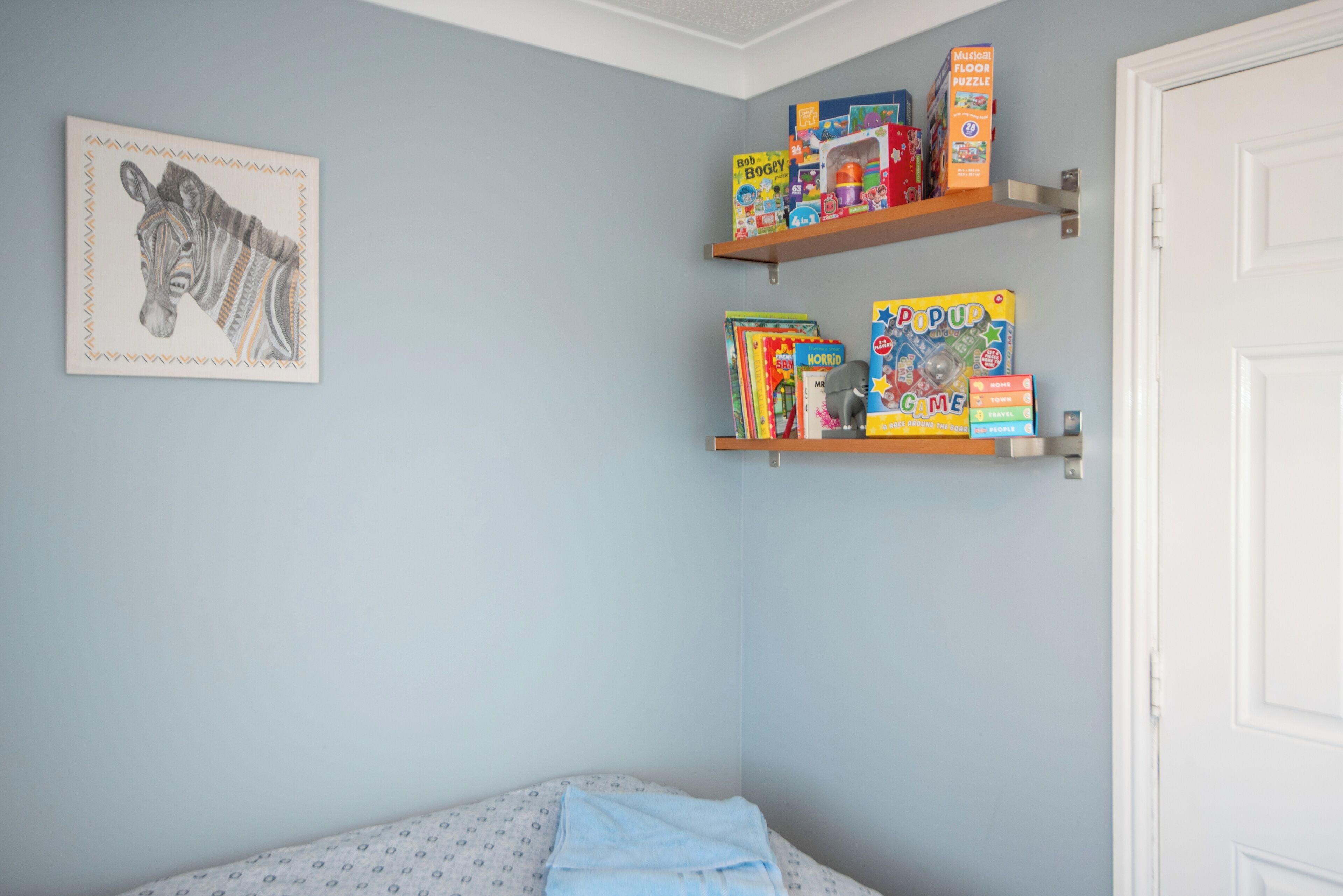 A bedroom with a bed, a wall-mounted zebra artwork, and two shelves with children's toys and books.