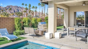 A backyard with a swimming pool, outdoor furniture, a grill, and a view of palm trees and mountains.