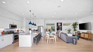 A modern kitchen with a central island, a dining area with a glass table, and a living room with a grey sofa and a TV.