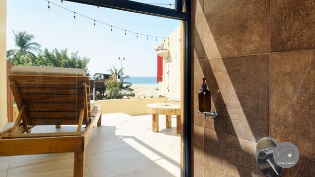A room with a view of the beach, a wooden chair, a small table, and a bottle on the wall. - Inigualable Suite, dos recamaras frente a Zicatela (Brisas de Zicatela)