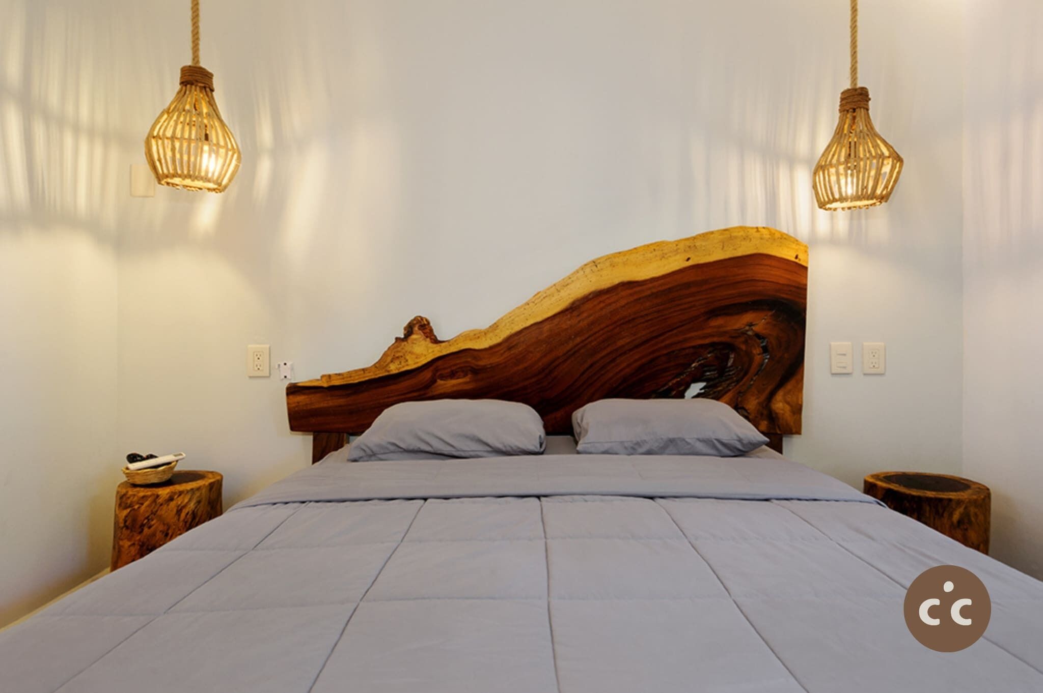 A bedroom with a wooden headboard, a bed with grey bedding, two bedside tables, and hanging light fixtures.
