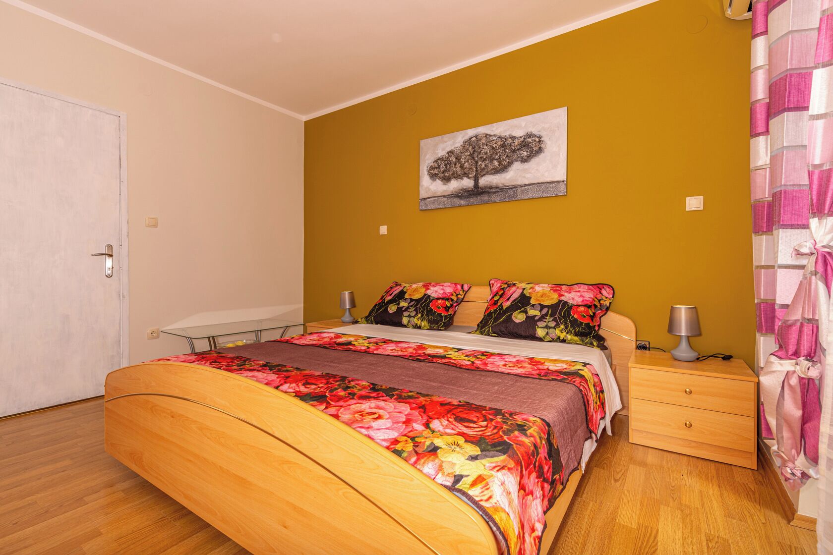A bedroom with a wooden bed, floral bedding, a nightstand with a lamp, and a painting of a tree on the wall.