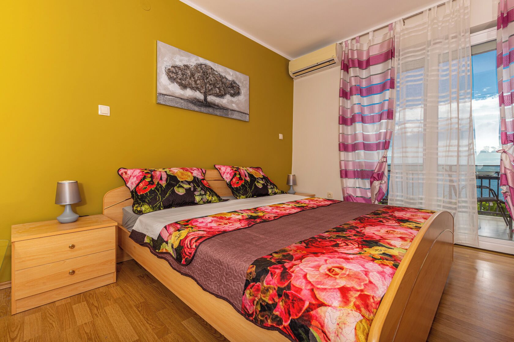 A bedroom with a bed, a nightstand, a lamp, a painting on the wall, and a view through the window.