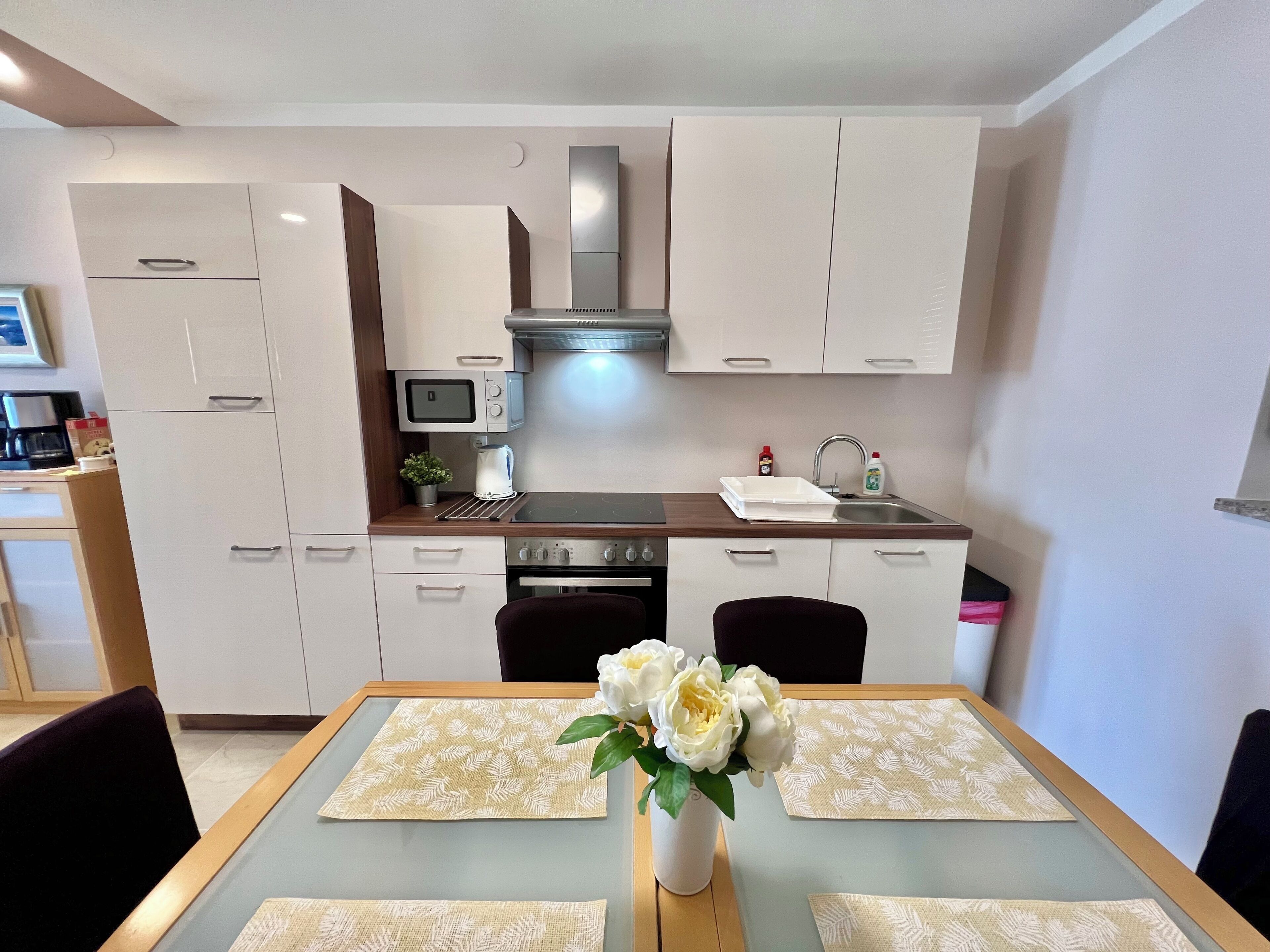 Una cocina moderna con armarios blancos, un microondas, una estufa y un lavamanos. Hay una mesa de comedor con un florero y manteles individuales.