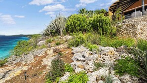 Paisatge costaner amb terreny rocos, cactus i un edifici amb mur de pedra i teulada de fusta.