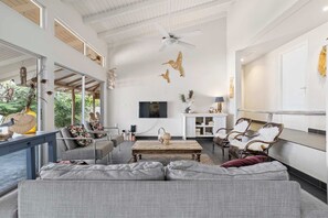 A spacious living area with a large grey sofa, a wooden coffee table, and wicker chairs. There is a ceiling fan, a TV mounted on the wall, and a window providing a view of greenery outside.