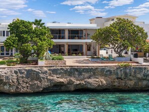 Une propriété située sur la côte, dotée d’une grande formation rocheuse, d’une eau turquoise cristalline et d’un bâtiment moderne à deux étages avec un espace extérieur aménagé pour s’asseoir.