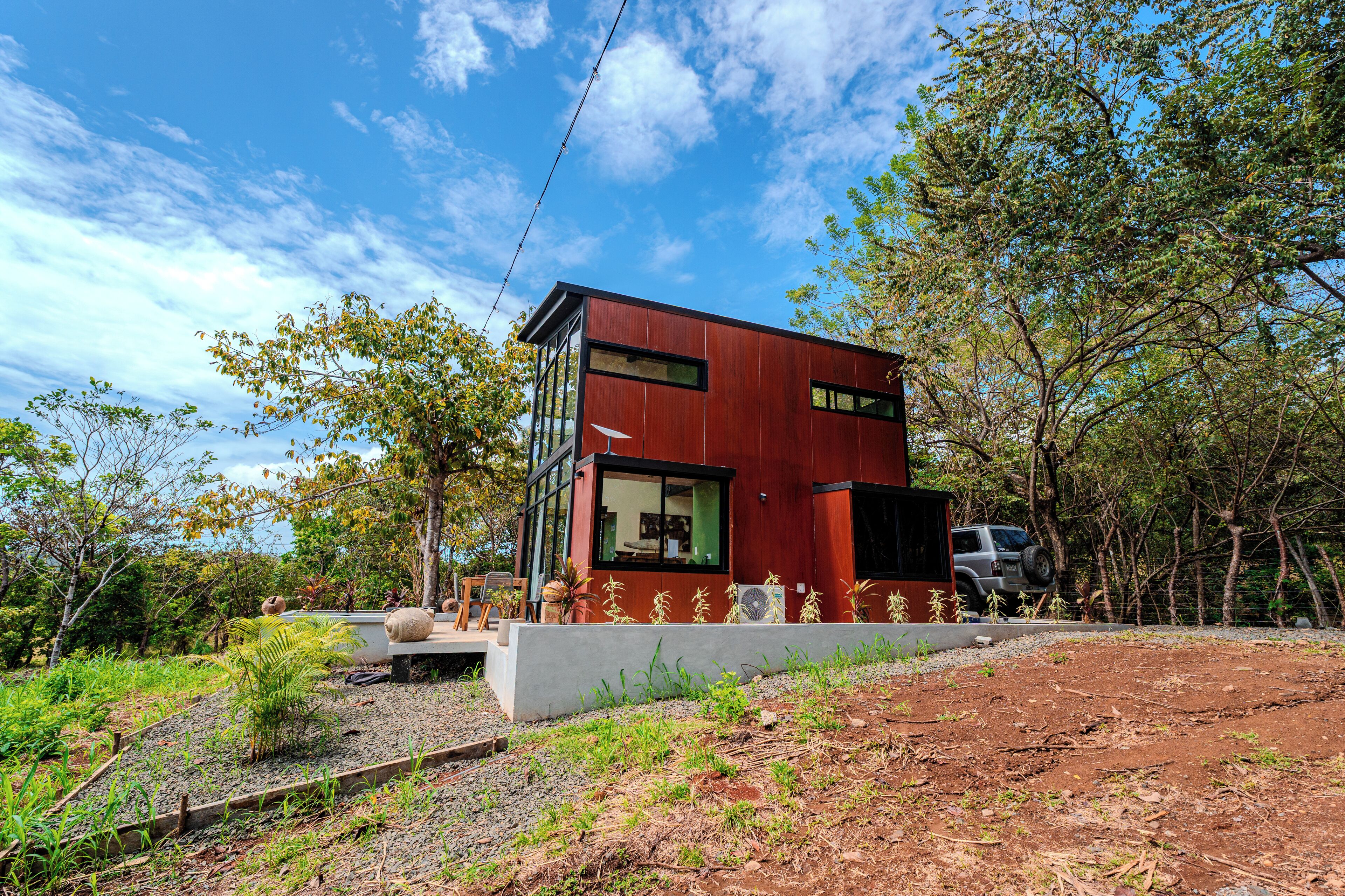 A modern two-story house with a red exterior, large windows, and a garden area with plants and a pathway.