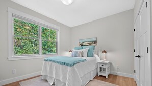 A bedroom with a large window, a bed with a blue and white bedspread, a nightstand with a lamp, and a painting on the wall.