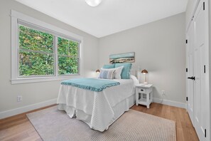 A bedroom with a large window, a bed with a blue and white bedspread, a nightstand with a lamp, and a painting on the wall. - TLC Cottage in Leland - New- sleeps 12! (Leland)