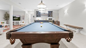A pool table with a ball in the center, a hanging light fixture, and a television in the background.