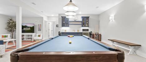 Ein Billardtisch mit einem Ball in der Mitte, eine hängende Leuchte und ein Fernseher im Hintergrund.