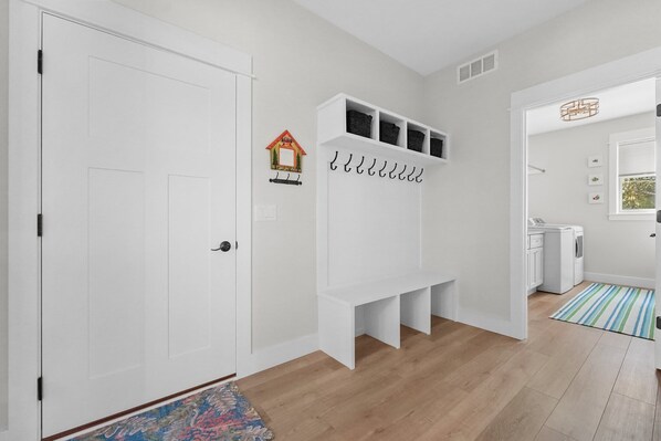 A hallway with a white door, a white bench with storage, a small decorative house, and a laundry area.