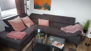 Ein modernes Wohnzimmer mit einem dunkelgrauen Sofa, rosa Kissen, einem Glastisch mit einer Zeitschrift, einer Wasserflasche und einer Topfpflanze.
