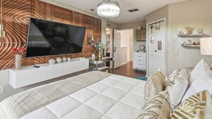 A modern bedroom with a large flat-screen TV, a bed with patterned bedding, a console table with decorative items, and a hallway leading to a bathroom.