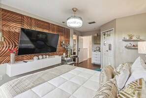 A modern bedroom with a large flat-screen TV, a bed with patterned bedding, a console table with decorative items, and a hallway leading to a bathroom.
