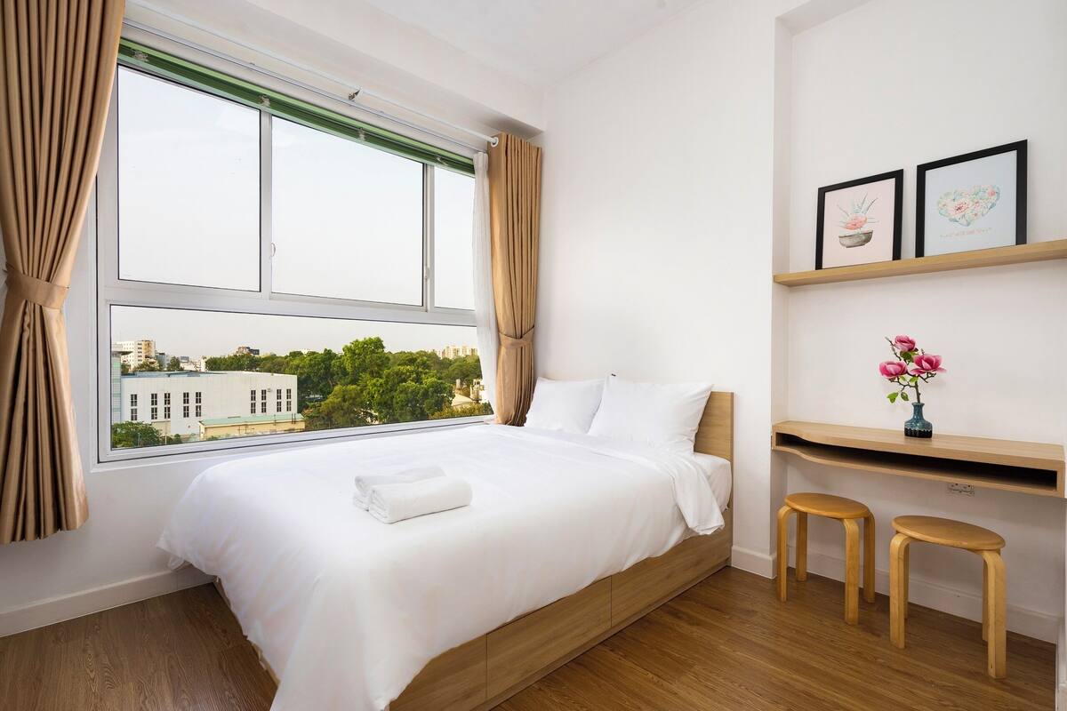 Ein ordentlich bezogenes Bett mit weißer Decke, ein Fenster mit Vorhängen und Blick auf Gebäude und Bäume.