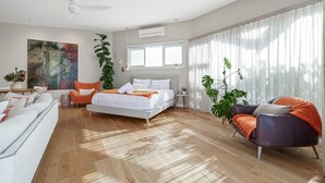 Un dormitorio moderno con cama, sofá y sillón. Se observa un ventanal con cortinas, un ventilador de techo y un cuadro en la pared.