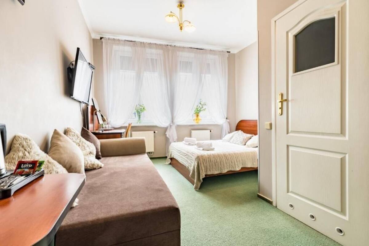 A hotel room with a bed, a sofa, a desk with a computer, and a window with sheer curtains.